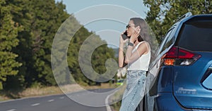 Young frustrated woman talking on mobile phone, leaning at her car with warning signals on