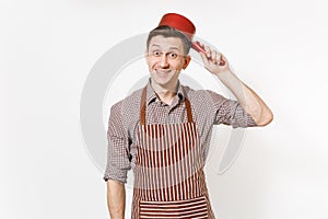Young fun crazy happy man chef or waiter in striped brown apron, shirt holding red empty stewpan on head isolated on