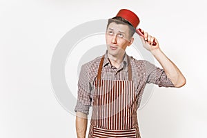 Young fun crazy happy man chef or waiter in striped brown apron, shirt holding red empty stewpan on head isolated on