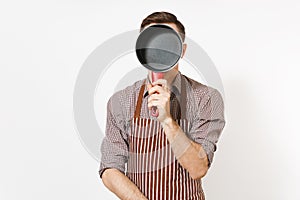 Young fun crazy man chef or waiter in striped brown apron, shirt holding red empty stewpan on face isolated on white