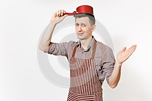 Young fun crazy sad man chef or waiter in striped brown apron, shirt holding red empty stewpan on head isolated on white