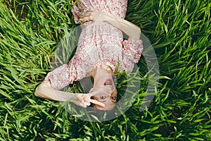 Young fun overjoyed beautiful woman in light patterned dress lying on grass showing victory sign resting in sunny