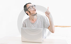 Young funny Asian man looking up and pointing finger up to present an empty space while using laptop computer in room. Korean or