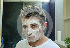 Young funny attractive man at home bathroom looking at toilet mirror applying facemask in his face finding himself weird and ugly