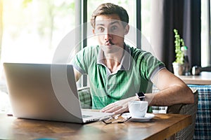 Young funny businessman in green t-shirt sitting and working on laptop and looking at camera with fish lips gesture crazy face