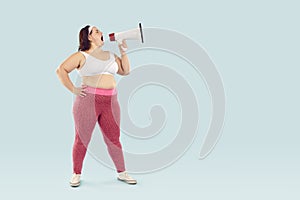 Young funny fat woman in sportswear screaming with mouthpiece on studio blue background.