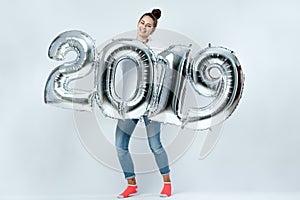 Young funny girl dressed in white t-shirt, jeans and pink socks holding balloons in the shape of numbers 2019 on the