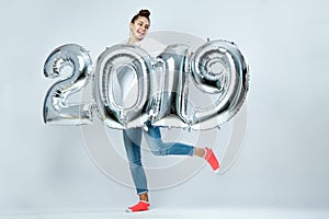 Young funny girl dressed in white t-shirt, jeans and pink socks holding balloons in the shape of numbers 2019 on the