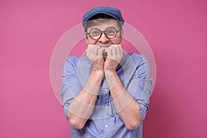 Young funny man in glasses and hat biting his nails on pink wall.