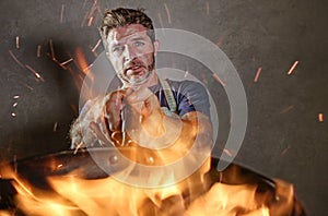 Young funny and messy home cook man with apron in shock holding pan in fire burning the food in kitchen disaster and domestic cook