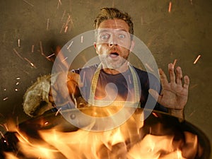 Young funny and messy home cook man with apron in shock holding pan in fire burning the food in kitchen disaster and domestic cook