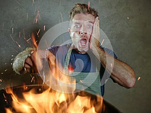 Young funny and messy home cook man with apron in shock holding pan in fire burning the food in kitchen disaster and domestic cook