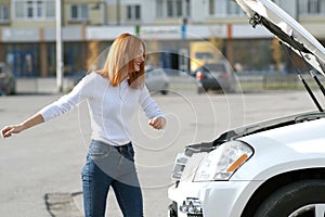 Young funny smiling woman driver near broken car with popped hood having a prbreakdown problem with her vehicle waiting for