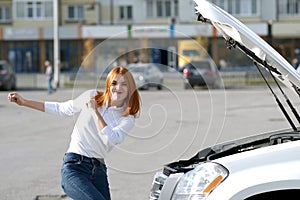 Young funny smiling woman driver near broken car with popped hood having a prbreakdown problem with her vehicle waiting for