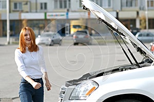 Young funny smiling woman driver near broken car with popped hood having a prbreakdown problem with her vehicle waiting for