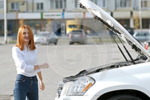 Young funny smiling woman driver near broken car with popped hood having a prbreakdown problem with her vehicle waiting for