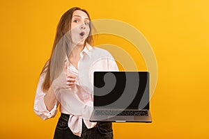 Young funny woman holds laptop with empty black screen workspace area show like gesture. Yellow background. Copy space, mock up