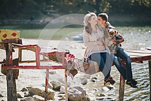 Young future parents and their dog in a funny costume sitting on a wooden bridge and having picnic near lake