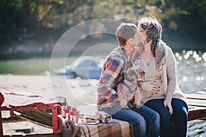 Young future parents and their dog in a funny costume sitting on a wooden bridge and having picnic near lake