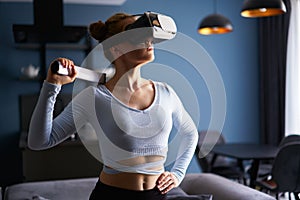 Young futuristic blonde girl wearing virtual reality headset, holding controller playing a video game at home. Woman