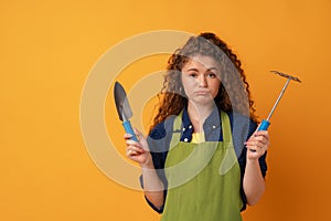 Young gardener woman wearing apron holding gardening tools against yellow background