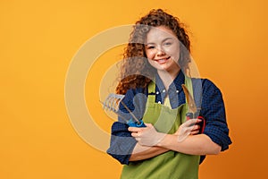 Young gardener woman wearing apron holding gardening tools against yellow background