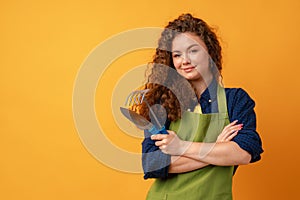 Young gardener woman wearing apron holding gardening tools against yellow background