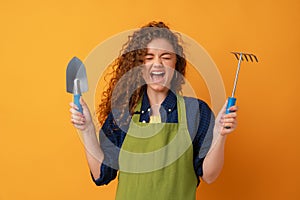 Young gardener woman wearing apron holding gardening tools against yellow background