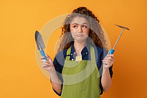 Young gardener woman wearing apron holding gardening tools against yellow background