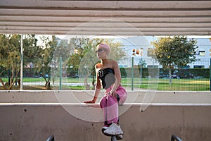 Young gay boy with pink hair and make-up and sunglasses is sitting on a wall resting, in the background a park with trees. Concept