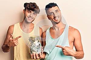 Young gay couple holding jar with savings smiling happy pointing with hand and finger