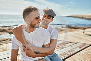 Young gay couple smiling happy sitting on the bench at the beach promenade