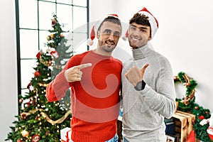 Young gay couple standing by christmas tree wearing hat pointing to the back behind with hand and thumbs up, smiling confident