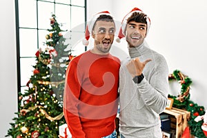 Young gay couple standing by christmas tree wearing hat smiling with happy face looking and pointing to the side with thumb up