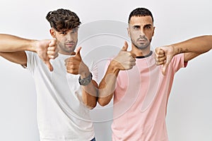 Young gay couple standing over isolated background doing thumbs up and down, disagreement and agreement expression