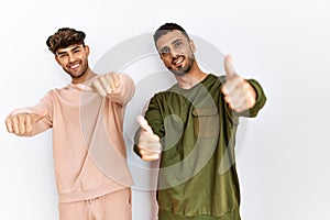 Young gay couple standing over isolated white background approving doing positive gesture with hand, thumbs up smiling and happy