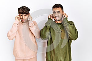 Young gay couple standing over isolated white background covering ears with fingers with annoyed expression for the noise of loud