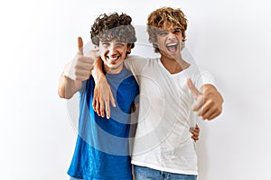 Young gay couple standing together over isolated background approving doing positive gesture with hand, thumbs up smiling and