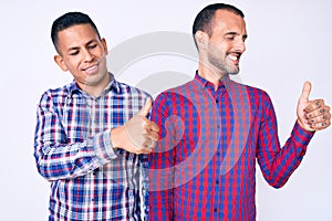 Young gay couple of two men wearing casual clothes looking proud, smiling doing thumbs up gesture to the side