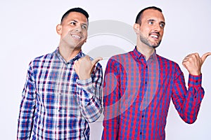 Young gay couple of two men wearing casual clothes smiling with happy face looking and pointing to the side with thumb up