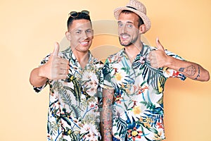 Young gay couple of two men wearing summer hat and hawaiian shirt doing happy thumbs up gesture with hand