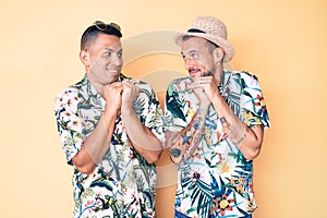 Young gay couple of two men wearing summer hat and hawaiian shirt laughing nervous and excited with hands on chin looking to the