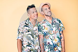 Young gay couple of two men wearing summer hat and hawaiian shirt looking away to side with smile on face, natural expression