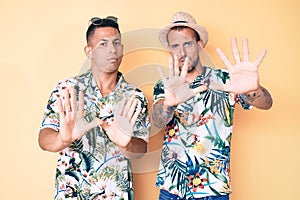 Young gay couple of two men wearing summer hat and hawaiian shirt moving away hands palms showing refusal and denial with afraid