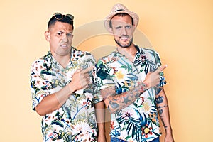 Young gay couple of two men wearing summer hat and hawaiian shirt pointing aside worried and nervous with both hands, concerned