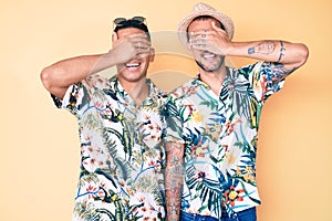 Young gay couple of two men wearing summer hat and hawaiian shirt smiling and laughing with hand on face covering eyes for