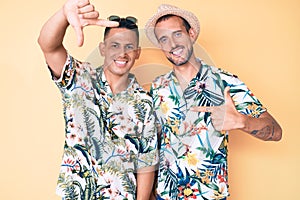 Young gay couple of two men wearing summer hat and hawaiian shirt smiling making frame with hands and fingers with happy face