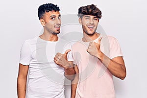 Young gay couple wearing casual clothes smiling with happy face looking and pointing to the side with thumb up