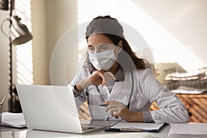 Young general practitioner in medical face mask working at laptop