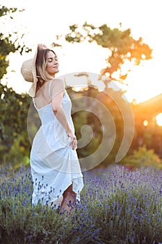Young Gentle Woman Lifts Hem of White Sundress and inhales Soothing Aroma of Lavender in Countryside